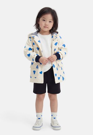 Fiatal gyermek bézs esőkabátban kék szívekkel, fehér pulóverben, fekete rövidnadrágban, fehér zokniban kék szívekkel és fehér sportcipőben áll.