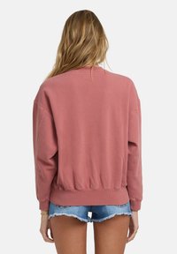 Voľný, skrátený sveter v prašnej ružovej farbe, s rebrovaným výstrihom a manžetami na rukávoch, v kombinácii s roztrhnutými džínsovými šortkami.
