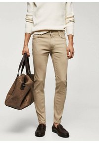 Jeans slim fit beige con costuras sutiles en los bolsillos, combinados con un suéter de punto beige y mocasines marrón oscuro. Una bolsa de viaje marrón completa el look.