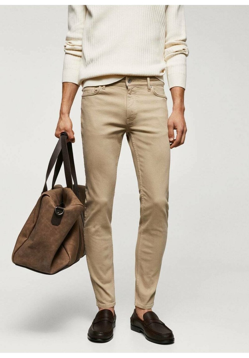 Jeans slim fit beige con costuras sutiles en los bolsillos, combinados con un suéter de punto beige y mocasines marrón oscuro. Una bolsa de viaje marrón completa el look.