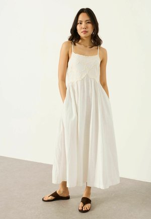 Mladá žena v bielych maxi šatách bez rukávov s kvetinovou výšivkou na vrchu, stojaca s rukami v vreckách pred jednoliatym pozadím.