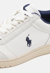 Vit lädersneaker med marinblå accenter, texturerad grå mockadetalj och kontrasterande vita snören. Har en marinblå broderad logotyp.