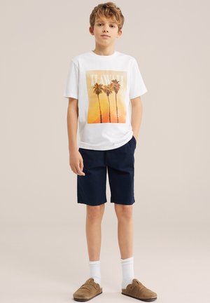Garçon aux cheveux châtain clair portant un T-shirt blanc avec un imprimé de palmier, un short bleu marine, des chaussettes blanches et des chaussures marron à enfiler, se tenant devant un fond beige.