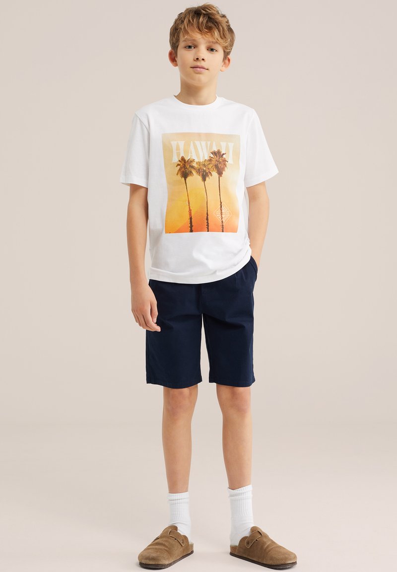 Dreng med lyst brunt hår iført en hvid T-shirt med palmebladsprint, marineblå shorts, hvide sokker og brune slip-on sko, stående mod en beige baggrund.