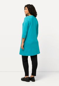 Türkise Tunika mit dreiviertel Ärmeln, runder Ausschnitt und geradem Schnitt; kombiniert mit schwarzen Slim-Fit-Hosen und schwarzen Schuhen.