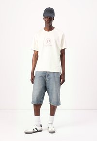 Hvid bomulds t-shirt med et cirkulært print og tekst, parret med lyseblå denimshorts, grå kasket og hvide sneakers med sorte striber.