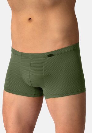 Männlicher Oberkörper trägt grüne enganliegende Boxershorts mit horizontalen Streifen vor einem einfarbigen hellen Hintergrund.