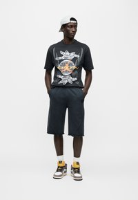 Μαύρο γραφικό μπλουζάκι με λευκό λεπτομερές τύπωμα, συνδυασμένο με αθλητικό σορτς σε σκούρο γκρι. Το μοντέλο φοράει ψηλά αθλητικά παπούτσια με κίτρινες και γκρι λεπτομέρειες.