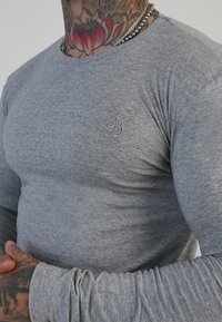 Camisa de manga larga gris, hecha de material suave, con un sutil logo embossado en el pecho y un diseño ajustado con mangas texturizadas.