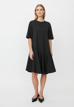 Kobieta stoi w czarnej sukience z krótkim rękawem, rozkloszowaną spódnicą i czarnych, spiczastych butach na tle jednolitego białego tła.