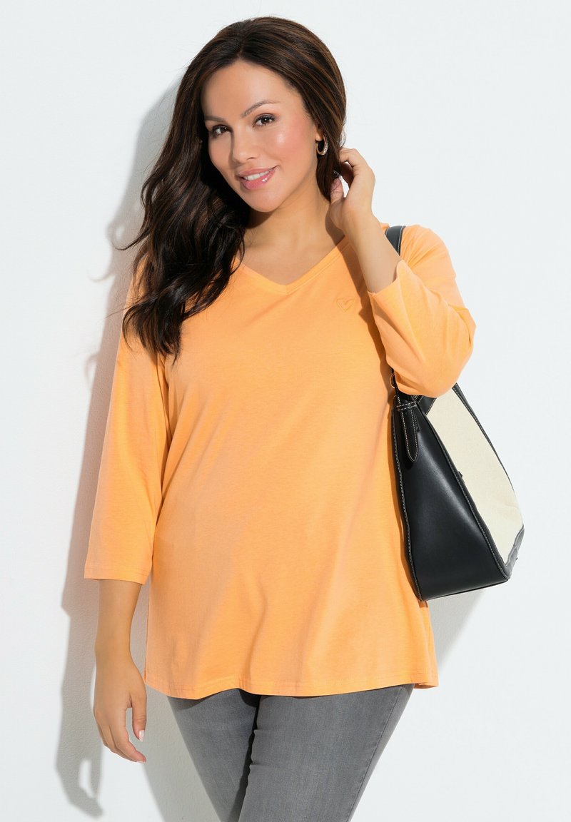 Haut orange à col en V et manches longues, coupe ample, associé à un jean gris et un sac à main noir avec des accents crème.