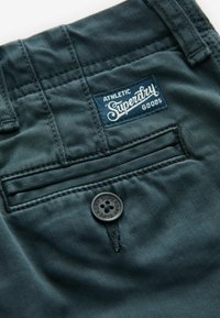 Mörkblått tyg med en strukturerad yta, med en bakficka och en rund svart knapp präglad med "Superdry." Inkluderar en blå etikett.