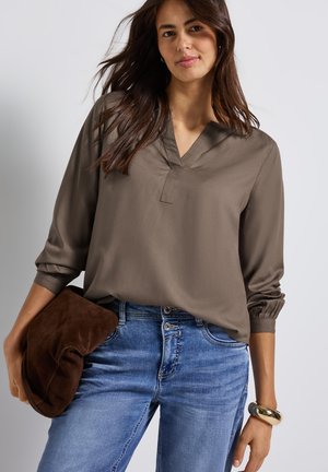 Brown, loose-fitting blouse with a V-neck and gathered sleeves, paired with blue jeans. The model holds a brown suede clutch.