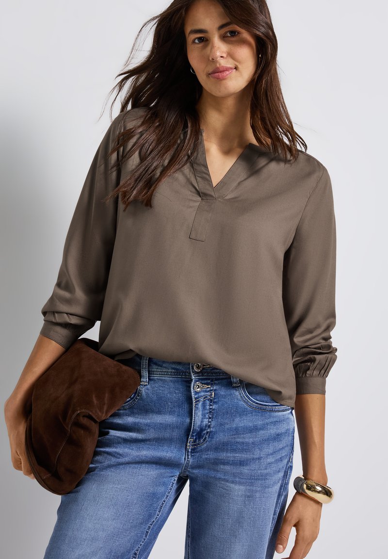 Blouse marron, ample avec un col en V et des manches froncées, associée à un jean bleu. Le mannequin tient une pochette en daim marron.
