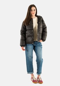 Giubbotto imbottito nero con cappuccio, con hardware dorato, abbinato a jeans azzurri arrotolati e scarpe da ginnastica bordeaux. Maglione beige strutturato sotto.