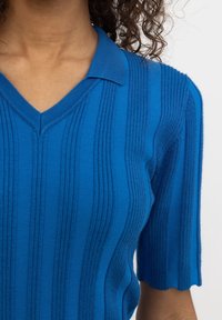 Top blu in maglia con scollo a V e maniche corte a ondina; presenta una texture a righe verticali per un aspetto strutturato.