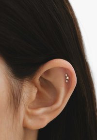 Pendiente de oro en forma de aro que presenta tres estrellas, con un diseño curvado, sujeto a la parte superior de la oreja con textura de piel visible y cabello oscuro al fondo.