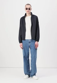 Jachetă neagră ușoară cu fermoar în față, purtată cu o cămașă crem și blugi largi de denim albastru. Modelul poartă ochelari de soare rotunzi și adidași albi.