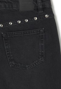 Vaqueros de mezclilla negra con un bolsillo trasero adornado con remaches plateados en la costura superior y una etiqueta de parche de cuero negro encima del bolsillo.