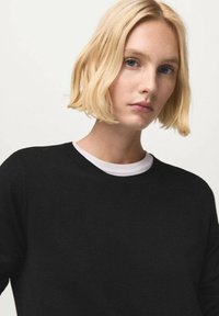 Schwarzer Pullover aus weichem Stoff mit rundem Halsausschnitt und weißem Innenkragen. Das Modell hat kurze blonde Haare und einen konzentrierten Gesichtsausdruck.