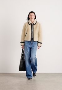 Leren shearling jas in crèmekleur met zwarte randen, wijde blauwe jeans en puntige zwarte schoenen, met een zwarte totebag. Effen achtergrond.