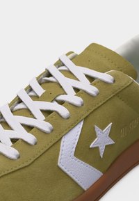 Παπούτσι αθλητικό από σουέτ σε ελαιδί πράσινο χρώμα με λευκά κορδόνια, λευκό αστέρι και λογότυπο σε σχήμα βέλους, και καφέ λαστιχένια σόλα, ορατό από το πλάι και από πάνω.