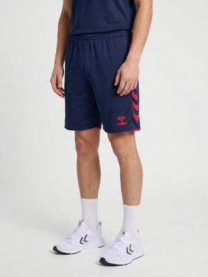 Mann trägt marineblaue Sportshorts mit rotem Logo und Pfeilstreifen, weiße Knöchelsocken und weiße Sportschuhe.