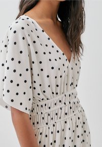 Lyhythihainen mekko kevyestä kankaasta, jossa on valkoinen tausta mustilla pilkuilla, syvä V-pääntie ja joustava vyötärö.