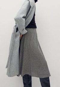 Persona con un abrigo gris con cinturón sobre una blusa negra, una falda midi de cuadros grises y botas altas negras contra un fondo liso.