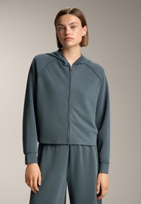 Felpa a zip color turchese chiaro con texture a coste, dotata di cappuccio, maniche raglan e orlo corto. Abbinata a pantaloni coordinati.