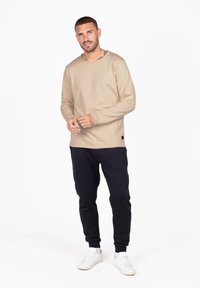 Beige Langarmshirt aus glattem Stoff, kombiniert mit schwarzen Jogginghosen. Weiße Sneakers runden das Outfit ab, das über ein dezentes Saumtag verfügt.