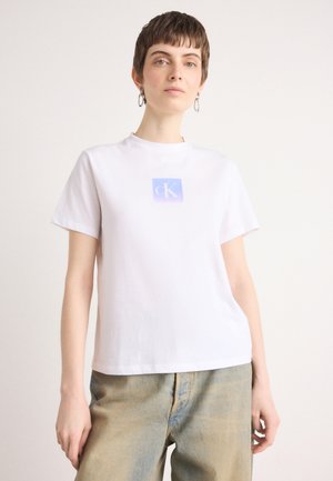 White cotton t-shirt with a round neckline and short sleeves. Features a purple and blue square logo in the center. Worn with light blue jeans.