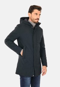Parka navy con cappuccio, chiusura con zip, bottoni a pressione, due tasche laterali e fodera interna testurizzata, indossato sopra un maglione grigio.