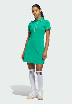 Grünes Polokleid mit Kragen, weißer Einfassung und Knopfleiste. Dazu kniehohe weiße Socken mit marineblauen Streifen und beigen Schuhen getragen.