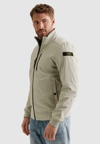 Mand iført lysebeige lynlåstrøje med sort lynlås og mærke på venstre overarm, kombineret med blå jeans, kigger til siden.