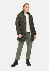 Olijfgroene pufferjack met een capuchon, met een gewatteerd ontwerp, een zilveren rits en zijzakken, gecombineerd met lichtgroene broek en zwarte laarzen.