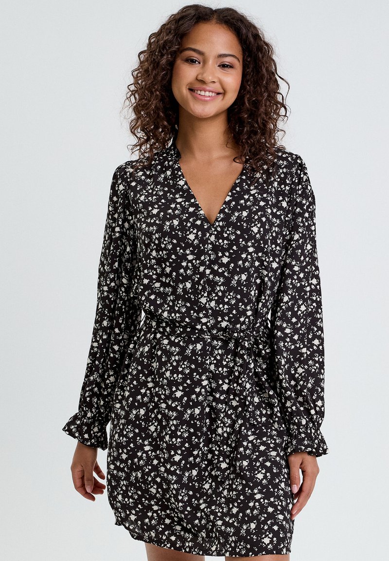 Robe noire avec imprimé floral blanc, col en V, manches longues et taille cintrée. Tissu doux avec une texture légère et fluide.