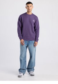Paarse sweatshirt met een ronde hals en geborduurd logo, gecombineerd met lichtblauwe jeans met rechte pijpen en witte sneakers.