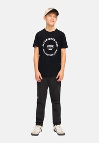 Camiseta negra con estampado gráfico blanco, jeans negros y gorra beige. Los zapatos son una mezcla de blanco y gris. Diseño simple y casual.