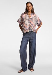Blusa a maniche corte con colletto, caratterizzata da un motivo geometrico colorato, abbinata a pantaloni di jeans scuri a gamba larga e sandali piatti.