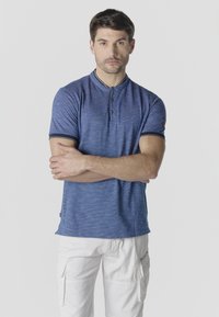 Homme portant un polo bleu à manches courtes avec col et un pantalon cargo blanc, debout les bras croisés devant un fond uni.