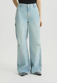 Vaqueros de pierna ancha de color azul claro, confeccionados en denim, con detalles desgastados y una cintura alta. Combinados con zapatos negros de punta con adornos.