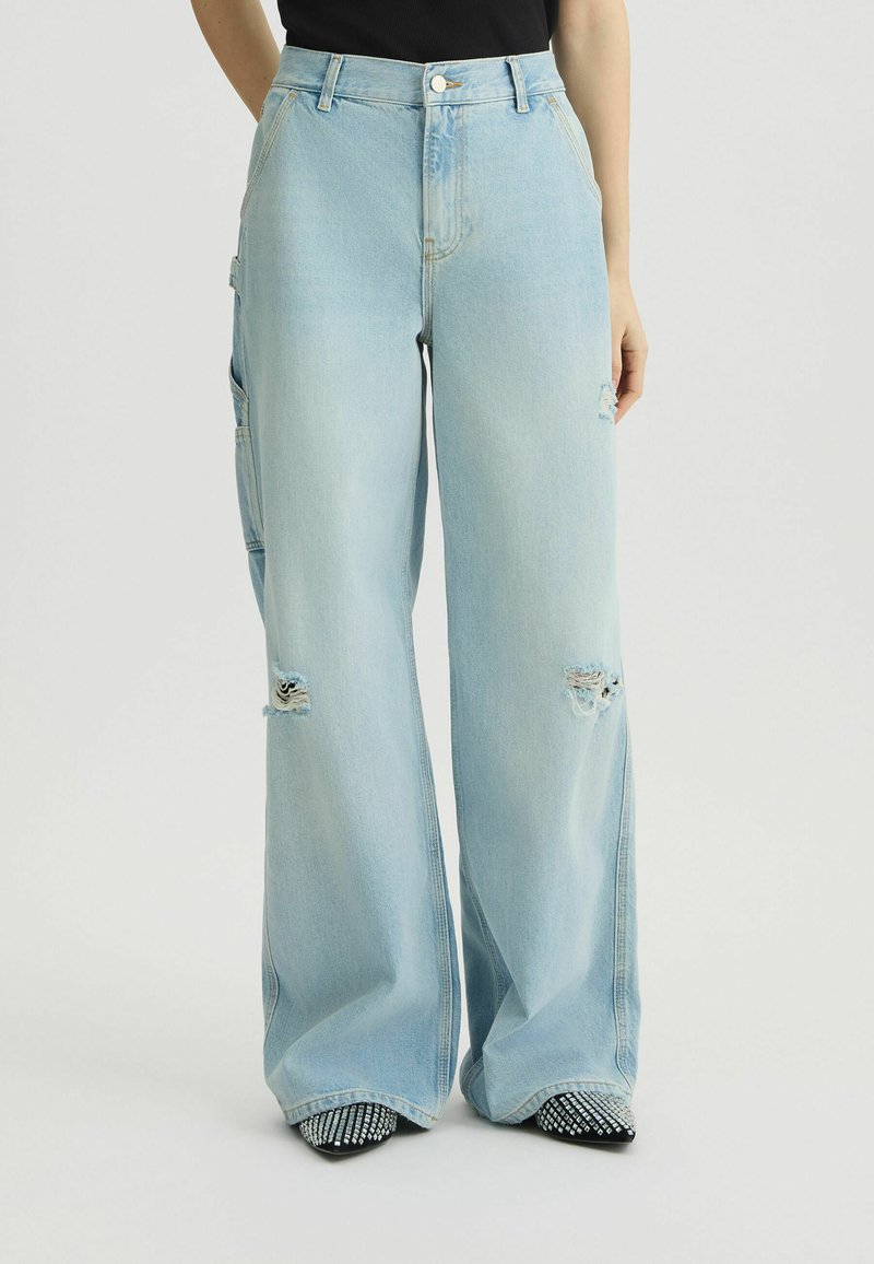 Vaqueros de pierna ancha de color azul claro, confeccionados en denim, con detalles desgastados y una cintura alta. Combinados con zapatos negros de punta con adornos.