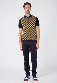 Knitted short-sleeve polo shirt featuring a gold and navy geometric pattern, paired with navy trousers and blue sneakers with red accents.