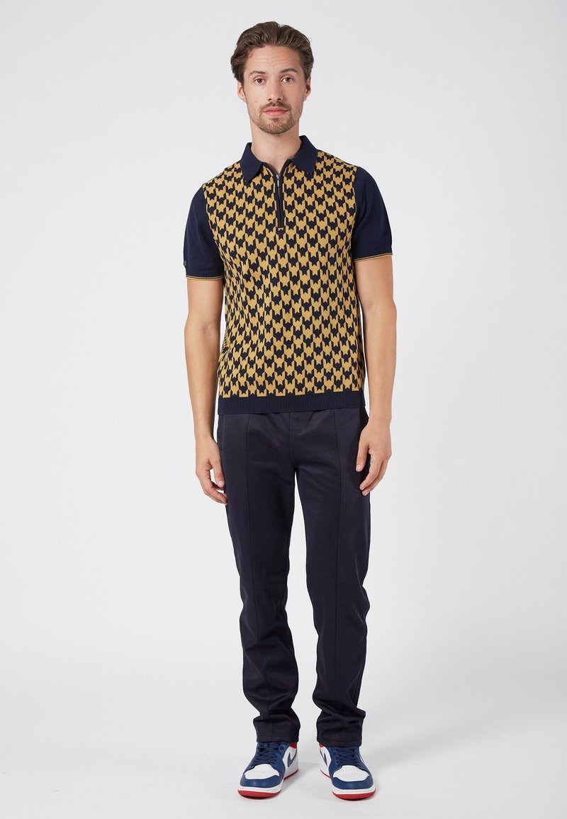 Knitted short-sleeve polo shirt featuring a gold and navy geometric pattern, paired with navy trousers and blue sneakers with red accents.