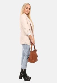 Femme aux longs cheveux blonds portant un blazer beige, un jean bleu clair, des bottes à talons noires, tenant un sac à main en cuir marron à ses côtés.