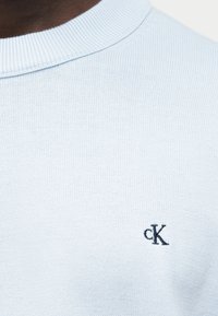 Κοντινό πλάνο του λαιμού ενός πλεκτού πουλόβερ ανοιχτού γαλάζιου χρώματος με μικρό μαύρο κεντημένο λογότυπο "cK" στο στήθος.