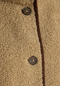 Beige strukturierter Stoffmantel mit einer flauschigen Außenschicht, ausgestattet mit dunkelbraunen Akzenten und einem großen Knopf, der mit "JOULES EST. 1868" beschriftet ist.