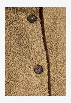 Beżowy płaszcz z teksturowanej tkaniny, z fuzowatą zewnętrzną warstwą, z ciemnobrązowymi akcentami i dużym guzikiem oznaczonym "JOULES EST. 1868."