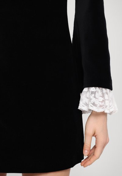 Svart fløyelskjole med lange ermer og hvite blondemansjetter. Stoffet er glatt med en rik tekstur, og designet er figurnært.